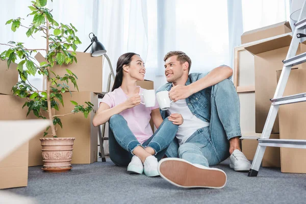 Усміхнена молода пара сидить на підлозі разом і смердить чашки з кавою, переходячи в новий будинок — стокове фото