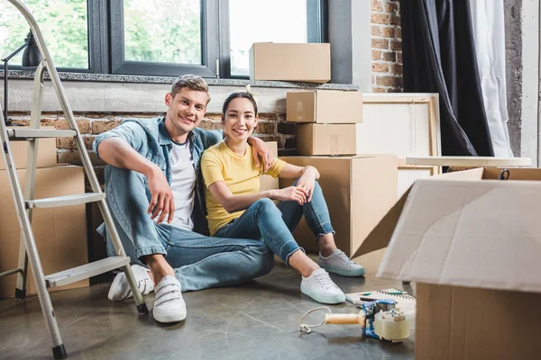 Sourire jeune couple assis sur le sol ensemble tout en déménageant dans une nouvelle maison — Photo de stock