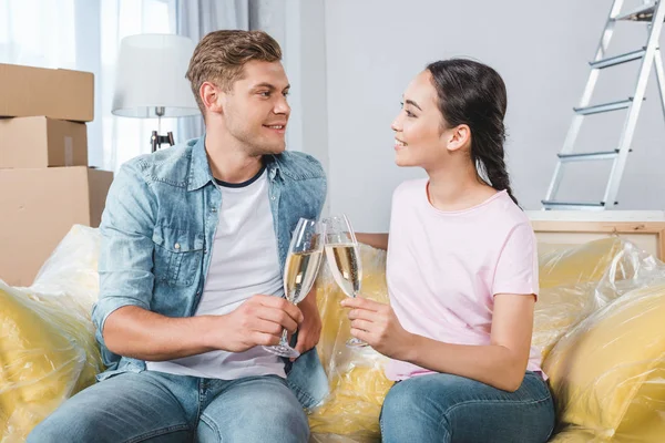Hermosa pareja joven tintineo copas de champán mientras está sentado en el sofá en el nuevo hogar - foto de stock