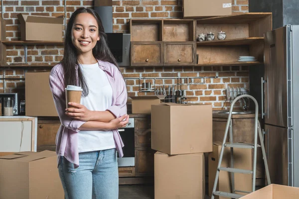 Giovane donna sorridente con tazza di carta di caffè e scatole in piedi sulla cucina di nuova casa — Foto stock