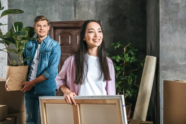 Beautiful young woman carrying canvas and her boyfriend holding ficus plant while moving into new home — Stock Photo