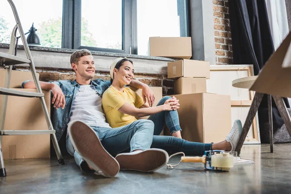 Hermosa pareja joven sentada en el suelo en una casa nueva rodeada de cajas - foto de stock