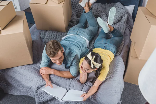 Vista de ángulo alto de feliz pareja joven con álbum de fotos tumbado en la cama mientras se muda a un nuevo hogar - foto de stock