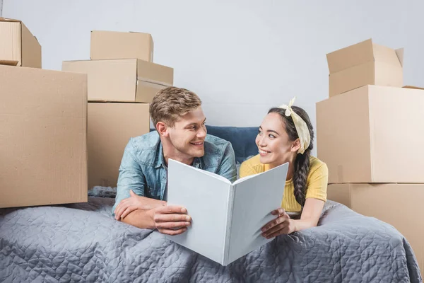 Feliz jovem casal com álbum de fotos deitado na cama enquanto se muda para uma nova casa — Fotografia de Stock