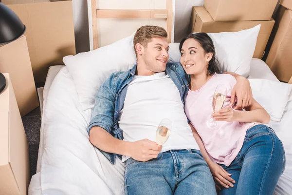 Vista de ángulo alto de la joven pareja acostada en la cama con copas de champán después de mudarse a un nuevo hogar - foto de stock