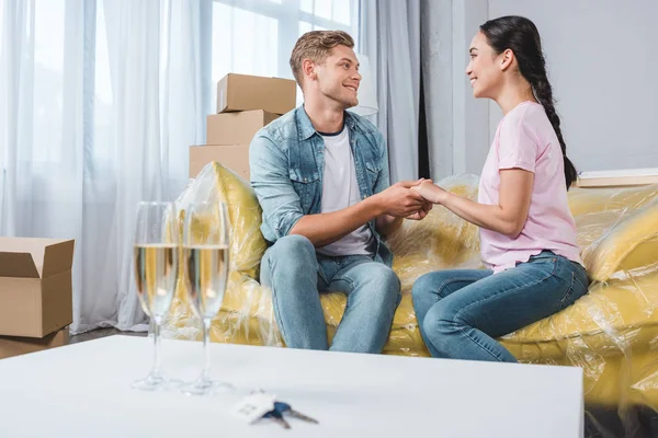 Hermosa pareja joven sentada en el sofá y tomados de la mano después de mudarse a un nuevo hogar con copas de champán en primer plano - foto de stock