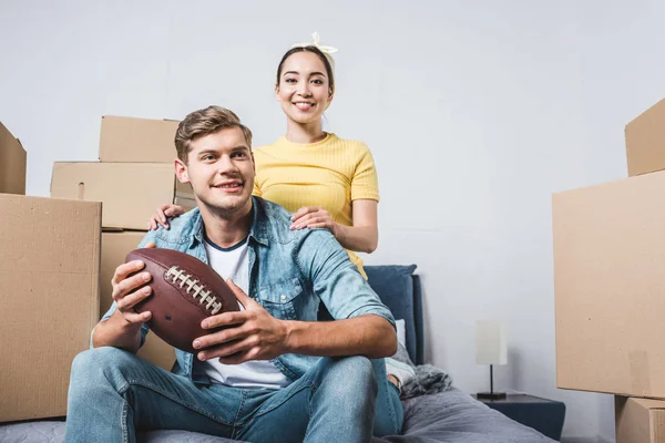 Jovem casal inter-racial sentado na cama com bola de futebol americano depois de se mudar para uma nova casa — Fotografia de Stock