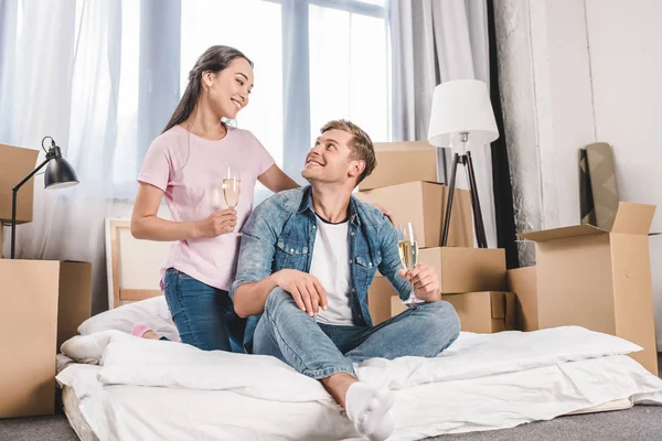 Hermosa pareja joven sentada en la cama con champán mientras se muda a un nuevo hogar - foto de stock
