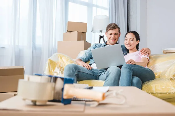 Hermosa pareja joven utilizando el ordenador portátil en el sofá mientras se muda a un nuevo hogar - foto de stock