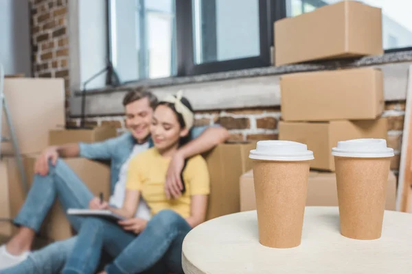 Beau jeune couple assis sur le sol tout en déménageant dans une nouvelle maison avec du café pour aller au premier plan — Photo de stock