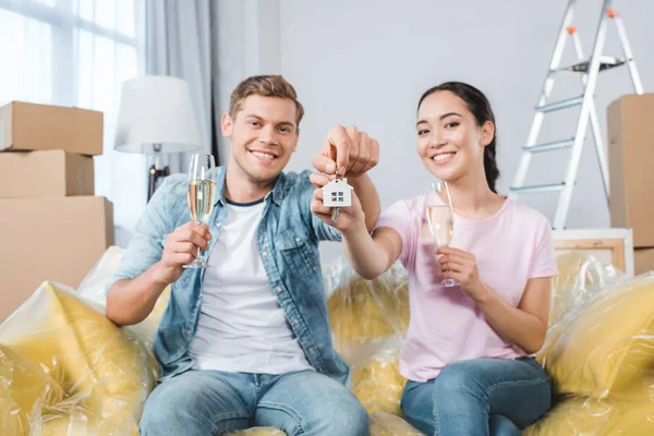 Щаслива молода пара в келихах шампанського і ключів сидить на дивані після переїзду в новий будинок — стокове фото