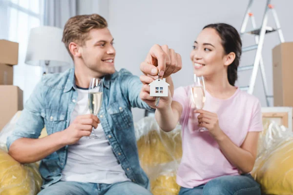 Beau jeune couple avec des verres de champagne et des clés assis sur le canapé après la réinstallation dans une nouvelle maison — Photo de stock