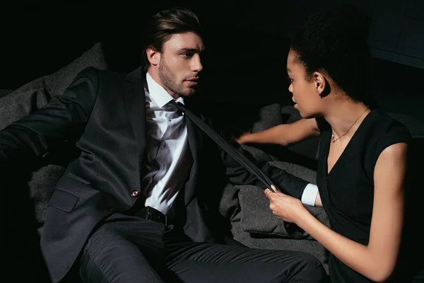 Young multiracial sexy couple in black clothing on sofa on dark background — Stock Photo