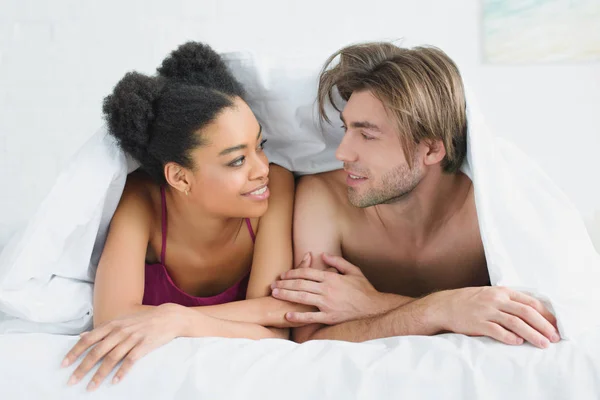 Portrait d'un jeune couple souriant et multiethnique amoureux couché sous une couverture au lit le matin — Photo de stock