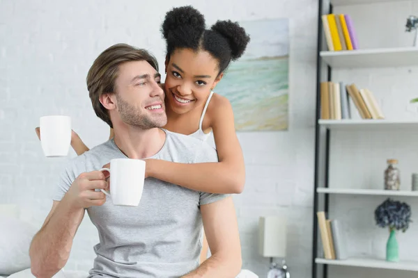 Retrato de casal multirracial sorridente com xícaras de café na cama de manhã — Fotografia de Stock