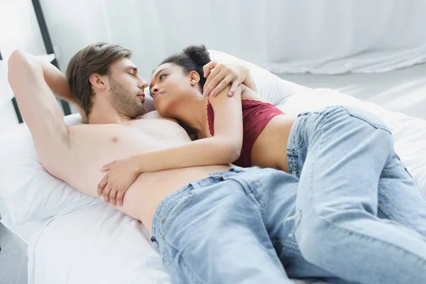Влюбленная многонациональная молодая пара, глядя друг на друга, лежа в постели вместе утром — стоковое фото