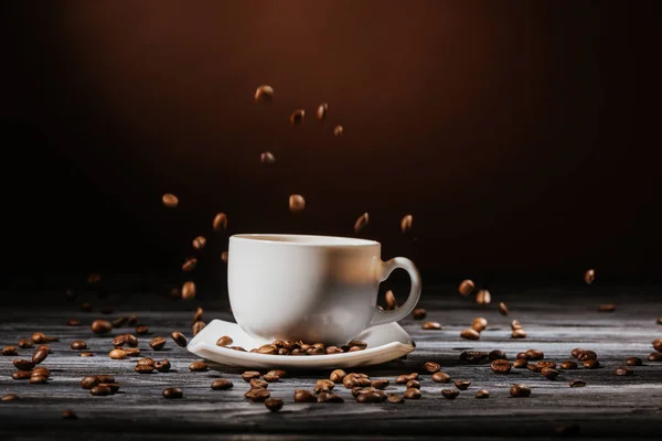 Primer plano de la taza con granos de café cayendo alrededor en la mesa de madera rústica - foto de stock