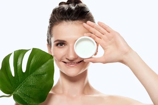 Hermosa mujer con crema facial en recipiente de plástico y hoja de monstera, aislado en blanco - foto de stock