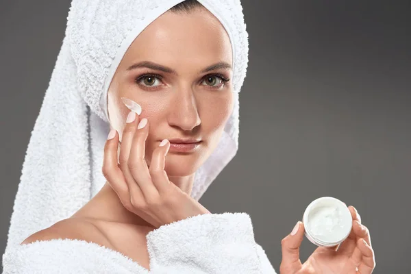 Hermosa chica en albornoz blanco y toalla aplicando crema cosmética en la cara, aislado en gris - foto de stock
