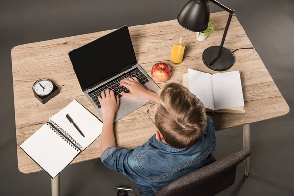 Vue aérienne de l'écolier faisant ses devoirs sur ordinateur portable à la table avec manuel, livre, plante, lampe, horloge, pomme et verre de jus — Photo de stock