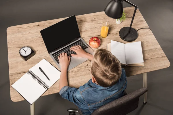 Overhead view of schoolboy doing homework on laptop at table with textbook, book, plant, lamp, clock, apple and glass of juice — Stock Photo