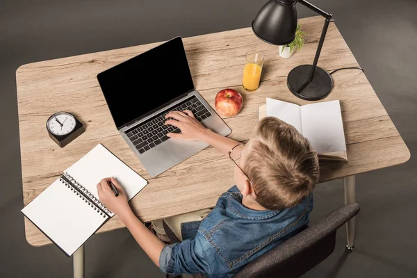 Elevated view of schoolboy doing homework on laptop at table with textbook, book, plant, lamp, clock, apple and glass of juice — Stock Photo