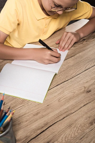 Partial view of schoolboy in eyeglasses writing in empty textbook at table with colour pencils — Stock Photo