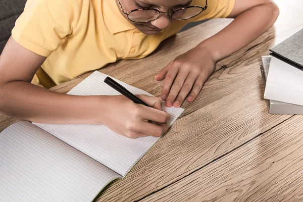 Vista parcial del colegial en anteojos escribiendo en libro de texto vacío en la mesa con pila de libros - foto de stock
