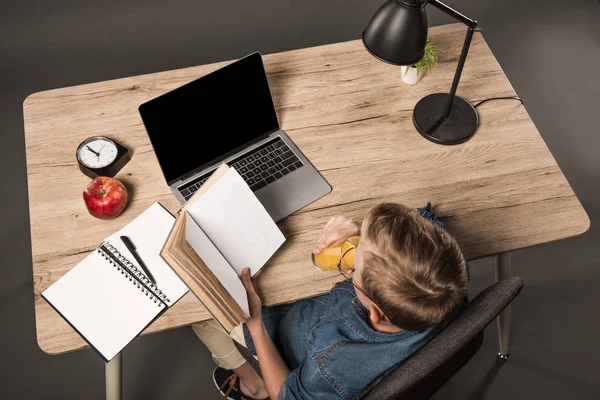 Вид зверху школяра читає книгу і п'є сік за столом з ноутбуком, підручником, лампою, рослиною, годинником і яблуком — стокове фото