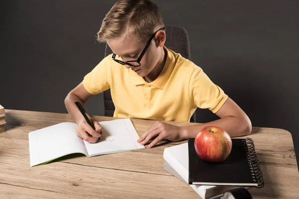 Focado menino da escola em óculos fazendo lição de casa na mesa com livros, livros didáticos e maçã em fundo cinza — Fotografia de Stock