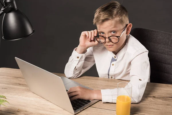 Serious little boy in eyeglasses looking at camera while sitting at table with laptop, lamp and glass of juice on grey background — Stock Photo