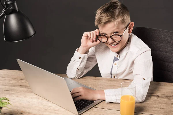 Улыбающийся мальчик в очках, смотрящий в камеру, сидя за столом с ноутбуком, лампой и стаканом сока на сером фоне — стоковое фото
