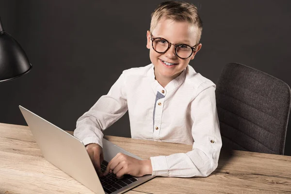 Happy little boy in eyeglasses looking at camera while sitting at table with laptop and lamp on grey background — Stock Photo