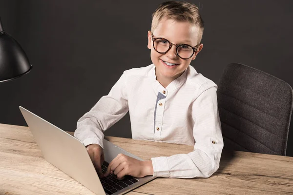 Heureux petit garçon dans des lunettes regardant caméra tout en étant assis à la table avec ordinateur portable et lampe sur fond gris — Photo de stock