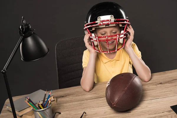 Estudante sorrindo colocando no capacete de futebol americano à mesa com bola, lâmpada, lápis de cor e livros sobre fundo cinza — Fotografia de Stock