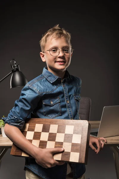 Улыбающийся маленький мальчик в очках держит шахматную доску рядом со столом с ноутбуком и лампой на сером фоне — стоковое фото