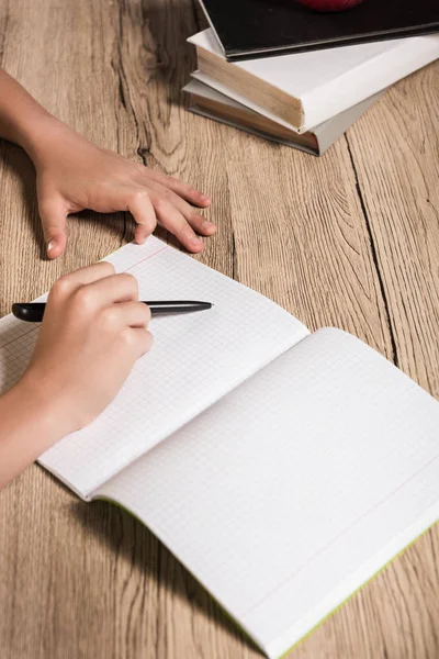 Image recadrée d'un écolier faisant ses devoirs dans un manuel vide à table avec une pile de livres — Photo de stock