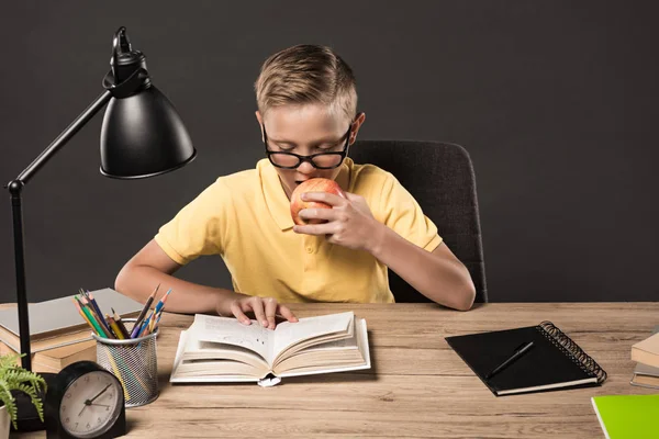 Scolaro in occhiali che mangia mela e legge libro a tavola con lampada, libri, orologio, matite a colori e libri di testo su sfondo grigio — Foto stock