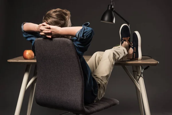 Rear view of schoolboy resting with legs on table on grey background — Stock Photo