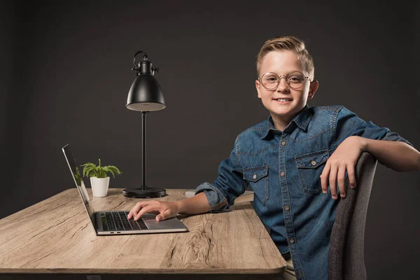 Усміхнений маленький хлопчик в окулярах використовує ноутбук за столом з рослиною і лампою на сірому фоні — стокове фото