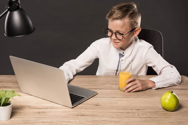Улыбающийся школьник в очках с помощью ноутбука за столом с лампой, стаканом сока, грушей и растением на сером фоне — стоковое фото