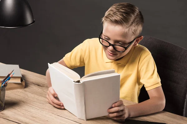 Colegial en anteojos leyendo libro en mesa con lámpara, lápices de colores y pila de libros sobre fondo gris - foto de stock