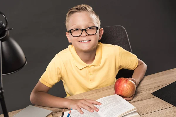 Lächelnder Schuljunge mit Brille, der in die Kamera blickt, während er am Tisch mit Apfel, Büchern und Lampe auf grauem Hintergrund sitzt — Stockfoto