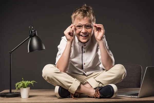 Улыбающийся маленький мальчик в очках сидит на столе с растением, лампой и ноутбуком на сером фоне — стоковое фото