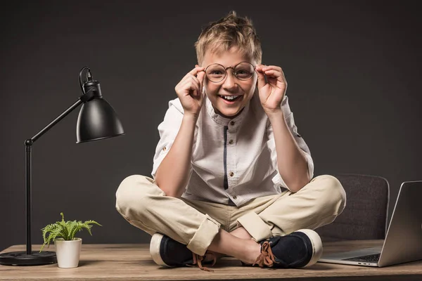 Niño sonriente en anteojos sentado en la mesa con la planta, la lámpara y el ordenador portátil sobre fondo gris - foto de stock