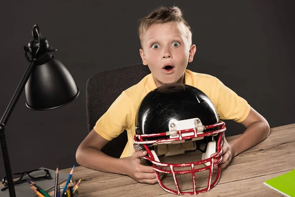 Shocked schoolboy sitting with american football helmet at table with eyeglasses, lamp, colour pencils and books on grey background — Stock Photo