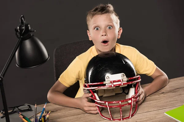 Colegial sorprendido sentado con casco de fútbol americano en la mesa con gafas, lámpara, lápices de color y libros sobre fondo gris - foto de stock