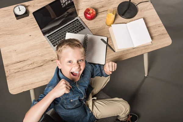 Overhead view of excited schoolboy gesturing by hands at table with laptop, book, textbook, apple, juice, lamp and clock — Stock Photo