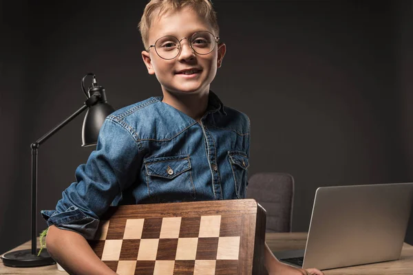Niño feliz en anteojos mirando a la cámara y sosteniendo tablero de ajedrez cerca de la mesa con el ordenador portátil y la lámpara sobre fondo gris - foto de stock