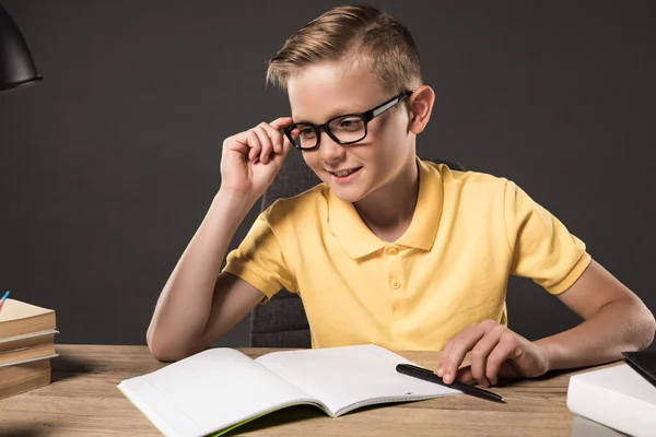 Colegial sonriente en gafas mirando hacia otro lado mientras hace la tarea en la mesa con pila de libros, libros de texto y lámpara sobre fondo gris - foto de stock